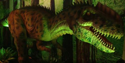 A large, realistic dinosaur model with green lighting, set against a forest backdrop.