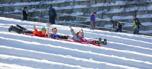 Three children sledding down a snowy hill, smiling and raising their arms in excitement. Snow-covered landscape in the background.