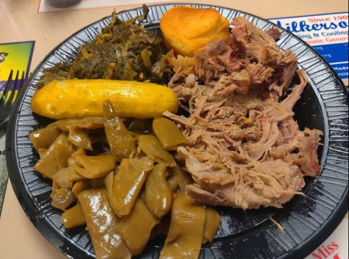 A black plate with pulled pork, collard greens, cornbread, yellow squash, and green beans.