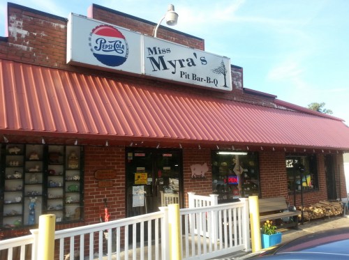 Exterior of Miss Myra's Pit Bar-B-Q, featuring a red metal roof, brick facade, and a sign with the restaurant's name.