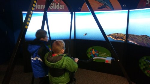 Two children interact with a flight simulator, looking at a large screen displaying a scenic ocean view.