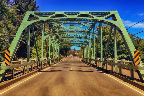 A green metal bridge spans a road, surrounded by trees under a clear blue sky.