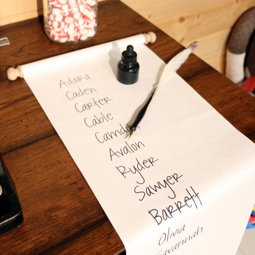 A wooden table with a scroll of paper listing names, a quill, and an ink bottle beside a jar of candy.