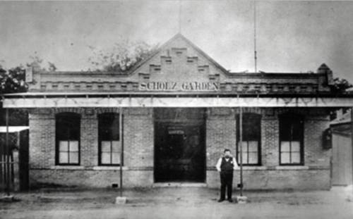 Historic black-and-white photo of Scholz Garden, featuring a brick building and a man in formal attire standing outside.