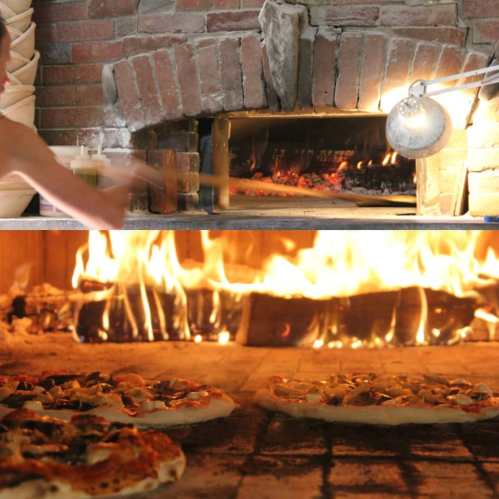 A wood-fired oven with pizzas baking inside, flames visible, and a hand using a pizza peel.
