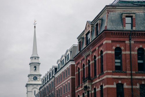 A tall church steeple rises above historic brick buildings under a cloudy sky.