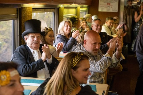 A lively group of people in a train car, some clapping, dressed in festive attire, enjoying a celebration.
