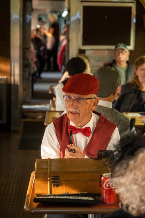 A man in a red cap and bow tie speaks into a microphone, seated at a table in a vintage train car.