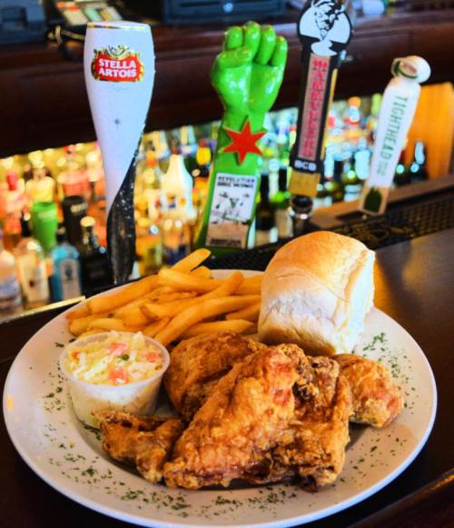 A plate of fried chicken with fries, coleslaw, and a bun, alongside beer taps in a bar setting.