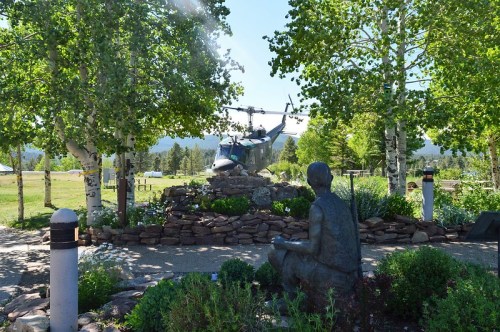 A statue sits in a garden with trees, overlooking a helicopter parked nearby on a sunny day.