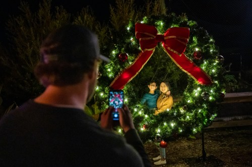 A person takes a photo of a couple posing inside a large, decorated Christmas wreath at night.