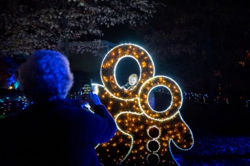 A person takes a photo of a child inside a glowing gingerbread decoration at a festive light display.