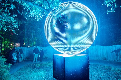 A glowing blue sphere made of lights stands in a park, with people walking nearby and trees in the background.
