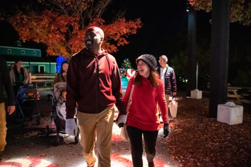 A couple walks hand in hand through a festive outdoor area, surrounded by colorful lights and autumn foliage.