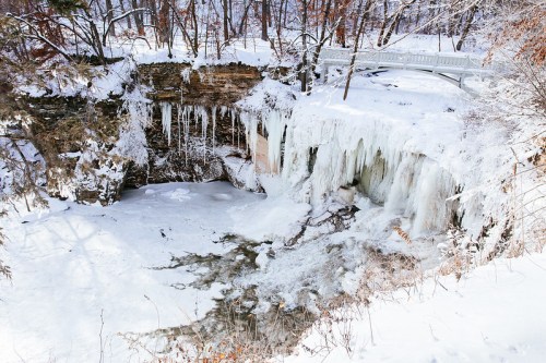 Frozen waterfall surrounded by snow-covered trees and a bridge in a winter landscape.