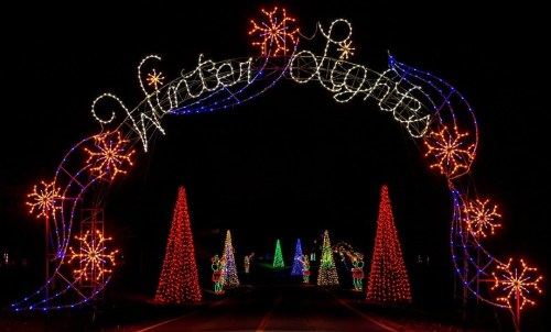 Colorful holiday lights form an archway reading "Winter Nights," with decorated trees and festive displays in the background.