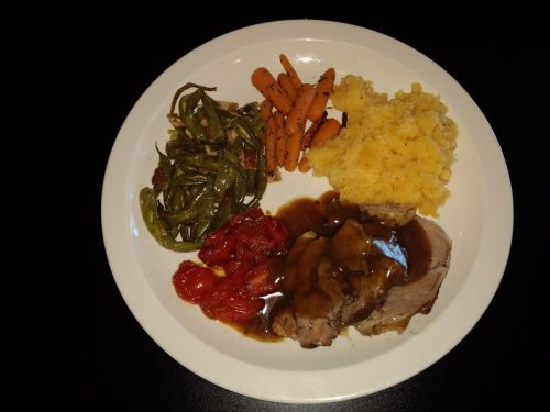 A plate of food featuring sliced roast beef with gravy, green beans, roasted carrots, mashed potatoes, and tomatoes.