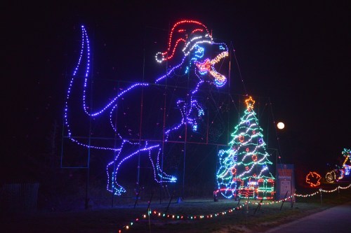 A large illuminated dinosaur wearing a Santa hat stands next to a colorful Christmas tree, set against a night sky.