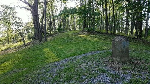 A grassy hill surrounded by trees, with a stone marker on the right and sunlight filtering through the leaves.