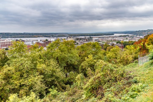 A scenic view of a river and cityscape surrounded by trees under a cloudy sky.
