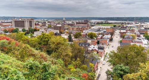 Aerial view of a cityscape with buildings, trees, and a river under a cloudy sky.