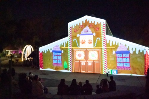 A building illuminated with colorful holiday projections, resembling a gingerbread house, with an audience seated in front.
