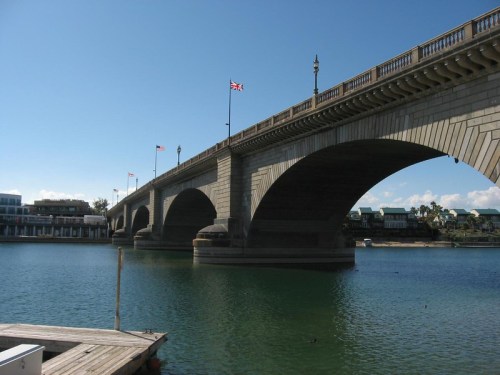 A stone bridge with arches spans a calm body of water, with flags flying above and buildings in the background.