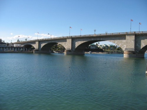 A stone bridge with arches spans a calm body of water, with flags flying and a clear blue sky above.