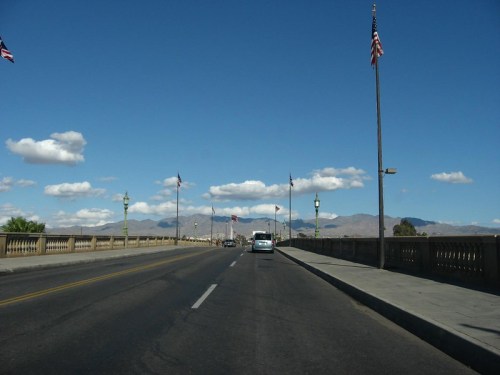 A view of a wide road on a bridge, lined with flags, under a blue sky with clouds and mountains in the background.