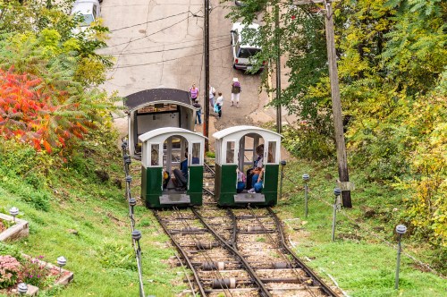 Two funicular cars on tracks, surrounded by greenery, with people walking nearby on a scenic hillside.