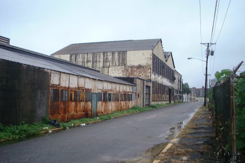 Abandoned industrial buildings along a wet, empty street, with overgrown grass and a cloudy sky.