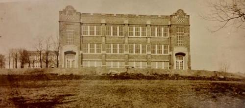 Historic brick building with large windows, set on a grassy hill, surrounded by bare trees. Black and white photo.