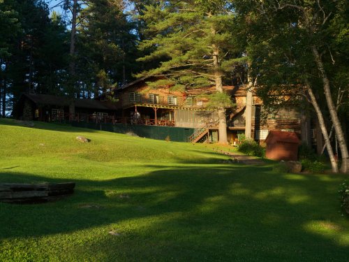 A rustic lodge surrounded by tall trees and a lush green lawn, bathed in warm evening light.