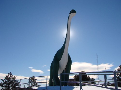 A large dinosaur statue stands against a bright blue sky, with the sun shining behind it, surrounded by snow and trees.