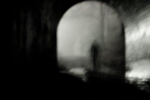 A blurred silhouette stands in a dark, foggy tunnel, creating an eerie and mysterious atmosphere.