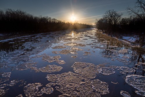 A serene river at sunset, with ice formations floating on the water and trees lining the banks.