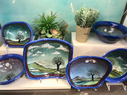 Colorful ceramic bowls and plates with landscape designs, featuring trees, mountains, and a night sky, displayed with plants.