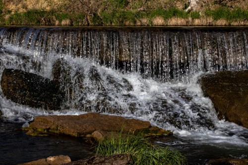A serene waterfall cascades over rocks, surrounded by lush green grass and a tranquil stream.
