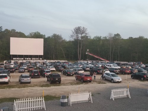 A view of Mendon Twin Drive-In with a large screen, filled parking lot, and trees in the background during twilight.