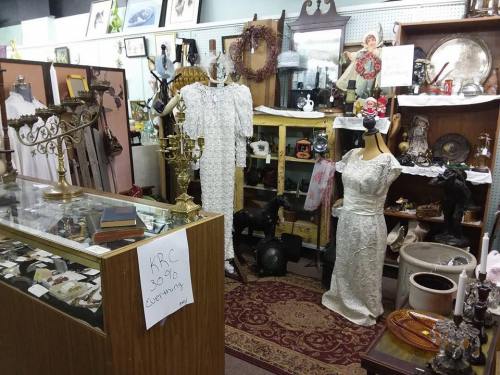 A vintage shop interior featuring antique dresses, decor, and various collectibles displayed on shelves and tables.