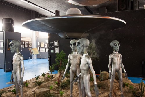 A display featuring a UFO and five alien figures in a museum setting, surrounded by plants and rocks.