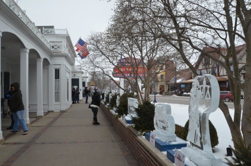 A snowy street scene with ice sculptures, people walking, and an American flag waving in the background.