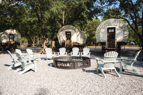 A cozy outdoor area with a fire pit surrounded by chairs and vintage-style wagons in a wooded setting.
