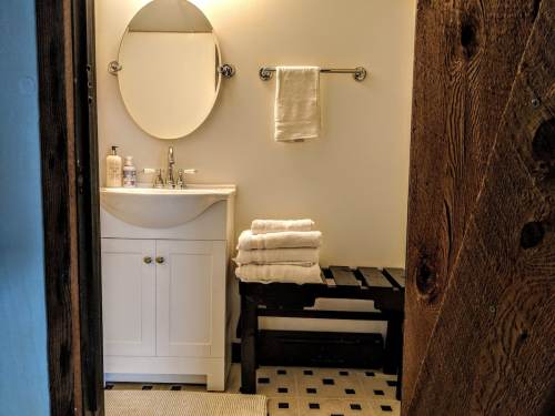 A small bathroom featuring a round mirror, sink, towel rack, and neatly stacked white towels on a wooden bench.