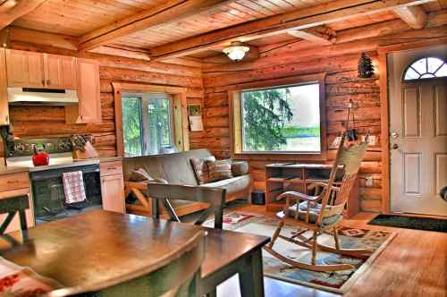 Cozy log cabin interior with a kitchen, living area, and rocking chair, featuring wooden walls and large windows.