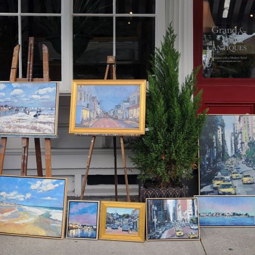 A display of various paintings on easels outside a shop, featuring landscapes and city scenes.
