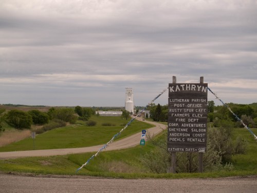 A rural road leads to Kathryn, featuring a sign for local businesses and a water tower in the background under a cloudy sky.