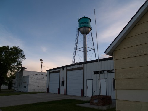 A water tower stands beside a fire station, with a clear sky in the background and a few buildings nearby.