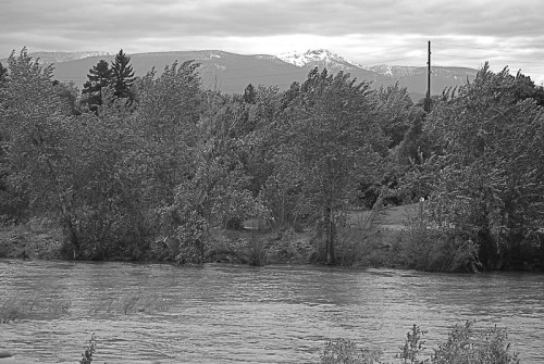Black and white landscape featuring a river, trees, and mountains in the background under a cloudy sky.
