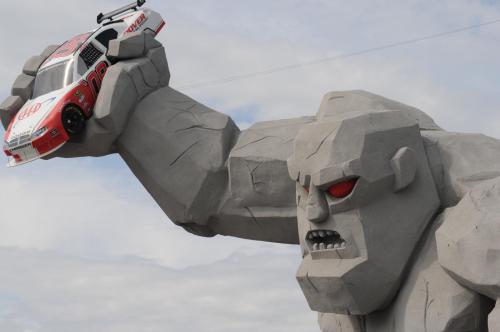 A giant stone figure with red eyes holds a toy race car in its fist against a cloudy sky backdrop.